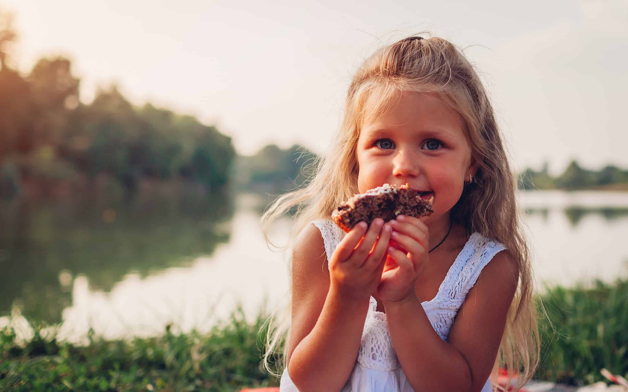 Little girl eating cake on family picnic by summer river. Child holding piece of pie