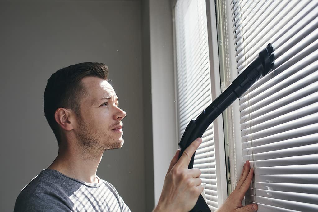 Man cleaning dust from window blind by vacuum cleaner at home