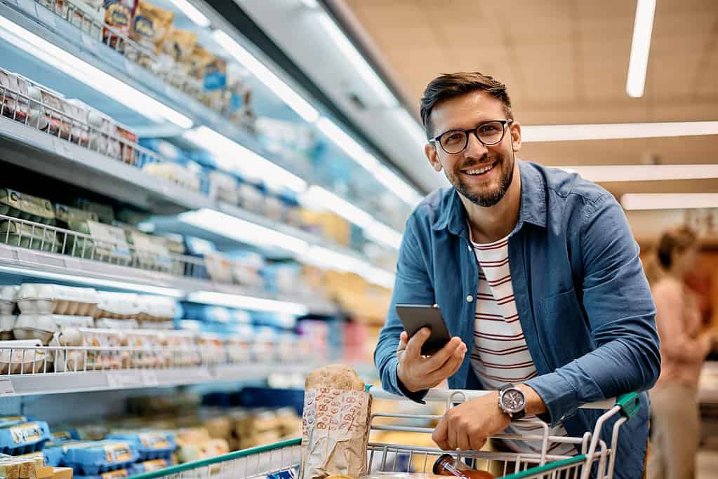 Happy man using mobile phone app while buying groceries in supermarket and looking at camera.