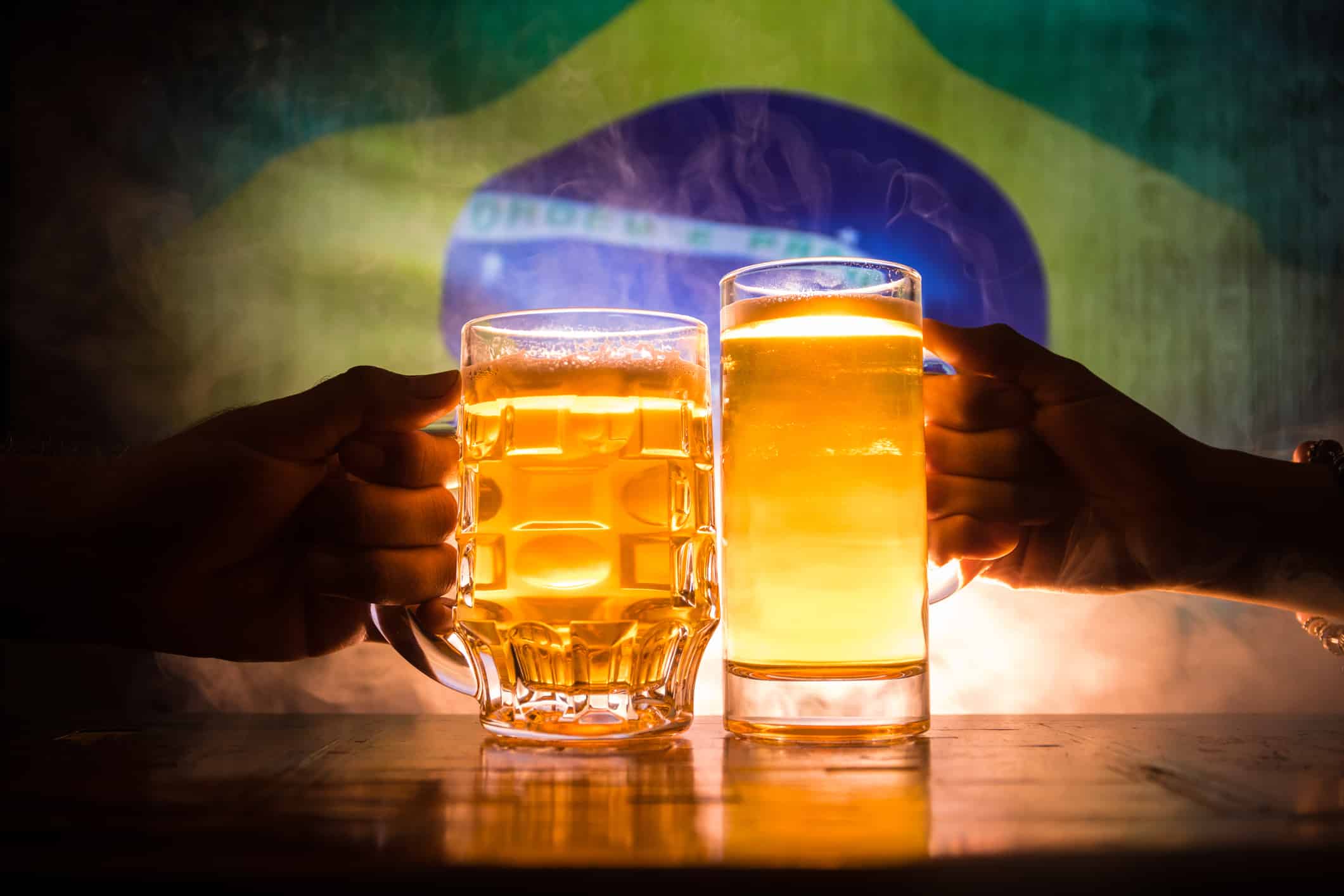 Two friends toasting (clinking) with glasses of light beer at the pub. Beautiful background with blurred view of flag of Brazil. Support your country with beer concept.