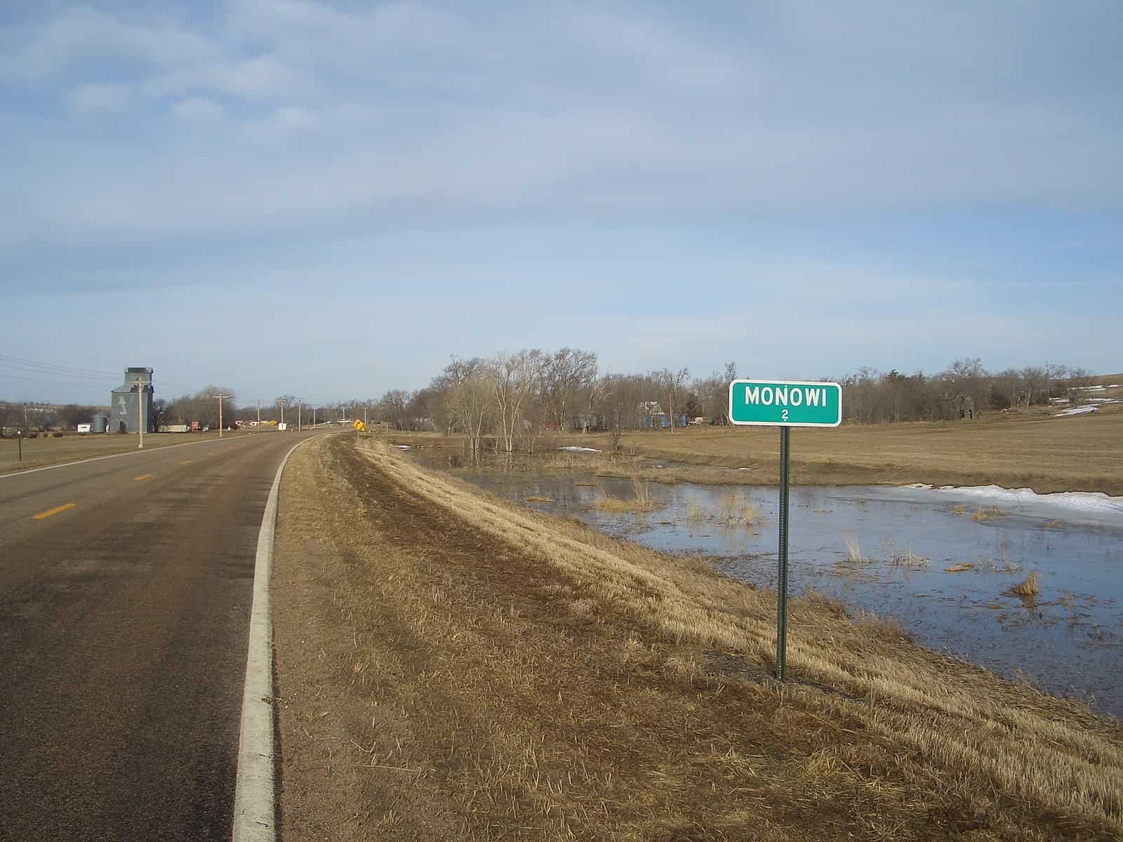 A sign that reads "Monowi" stands between a deserted road and a small pond.
