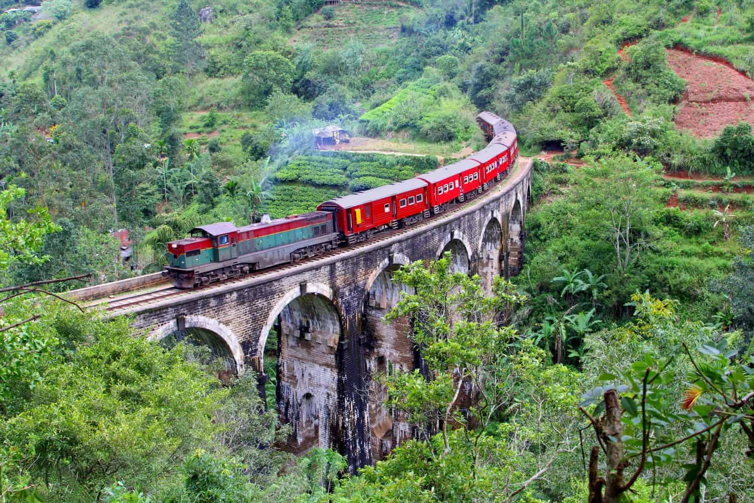 Red train with steam locomotive drives from Ella to Nuwara Eliya and Kandy over old Nine Arches bridge in jungle mountain landscapes. Uva Province, Sri Lanka