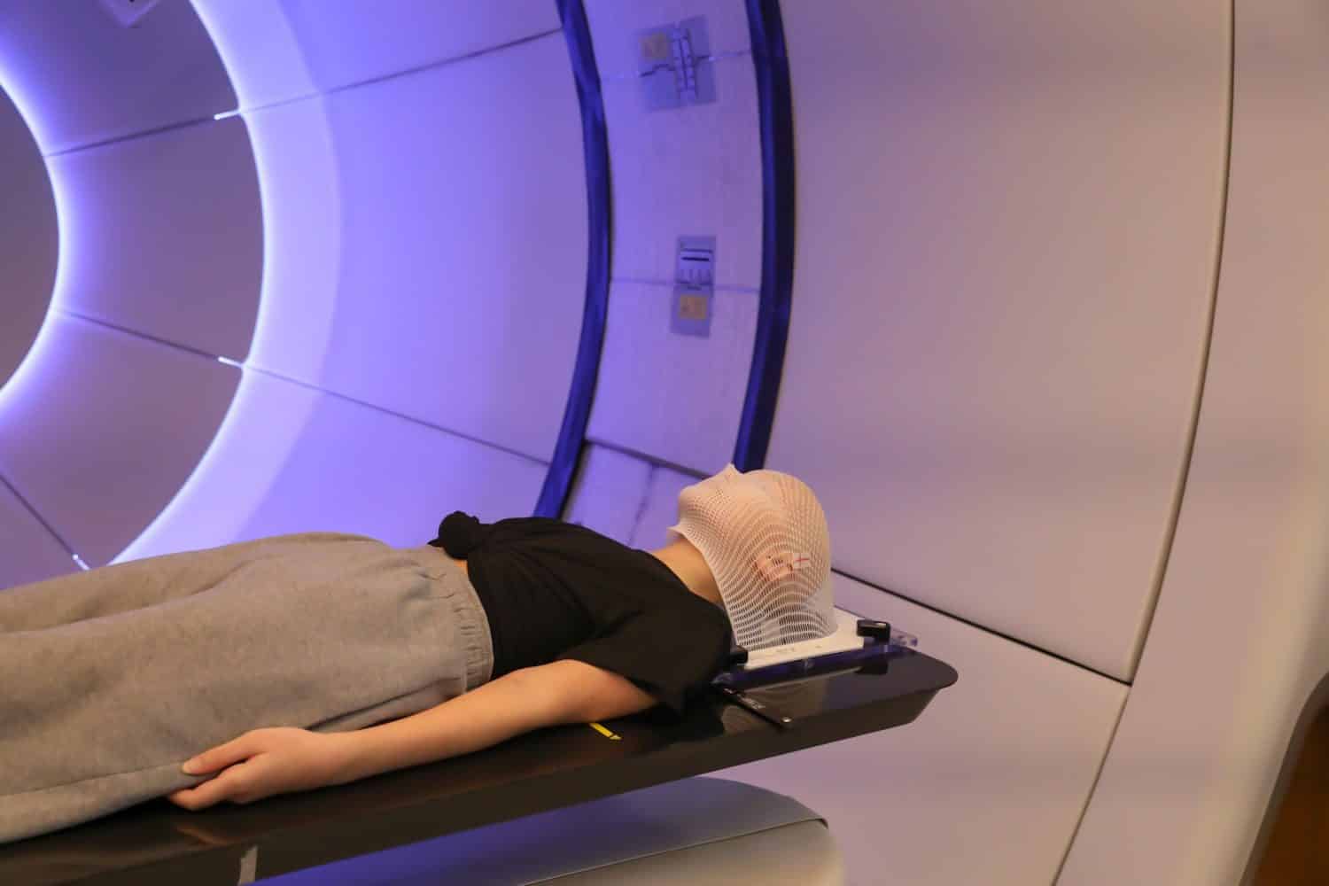 Radiation proton therapy. Brain cancer child patient lying on positioning bed with fixing mask on her head in treatment room.