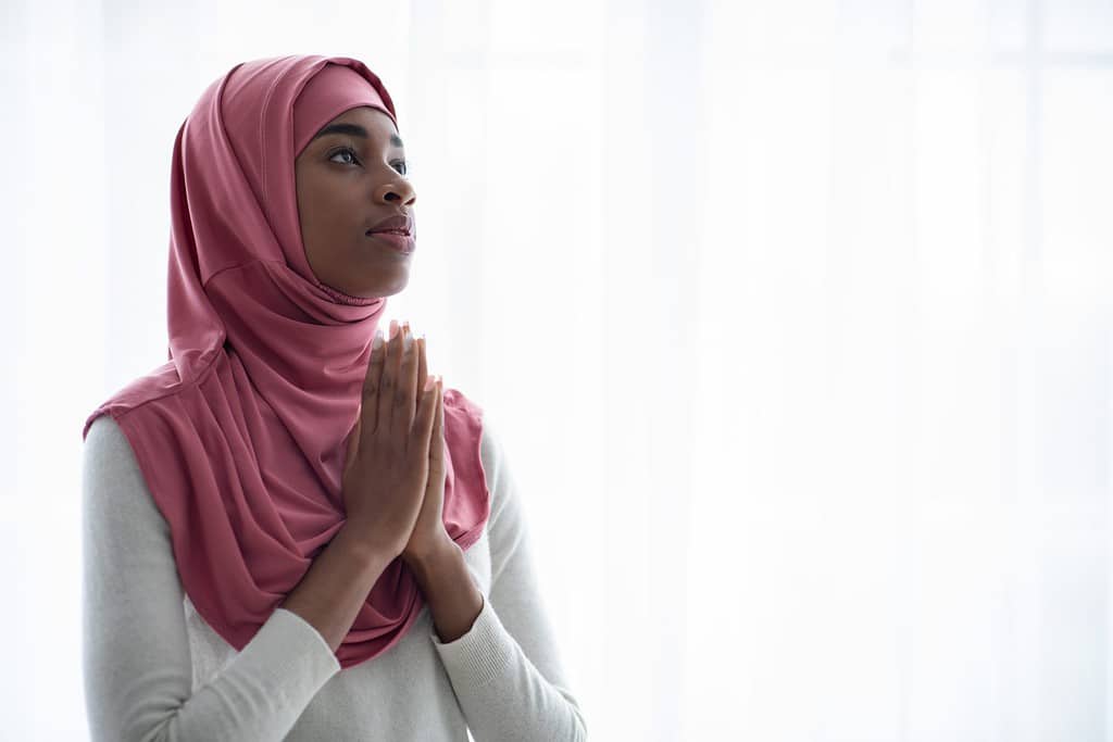 Young Black Muslim Woman In Pink Hijab Praying To Allah With Clasped Hands, Religious African Islamic Woman In Headscard Standing Near Window Over Daylight, Closeup Shot With Copy Space