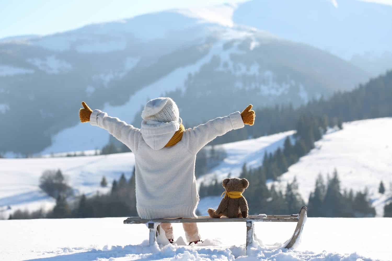 Сhild with toy teddy bear sits on a sled and looks at the winter snowy mountains.Winter family vacantion. Christmas celebration and winter holidays. Winter fun and outdoor activities with kids