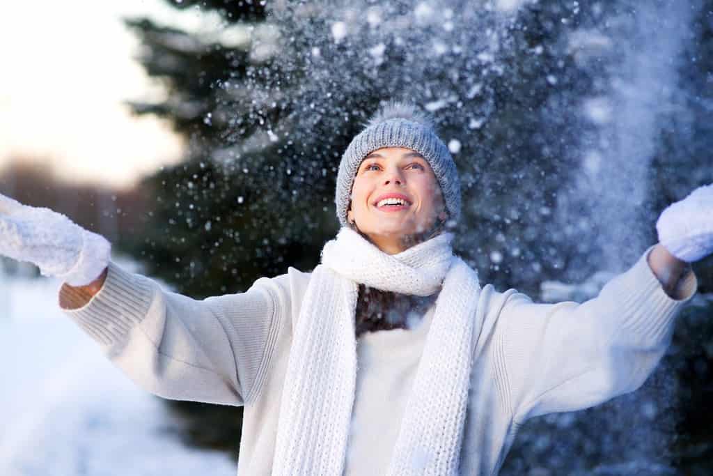 Portrait of happy beautiful girl, young joyful positive woman walking playing with snow, snowflakes, having fun outdoors in winter clothes, hat and scarf, smiling. Winter season, weather