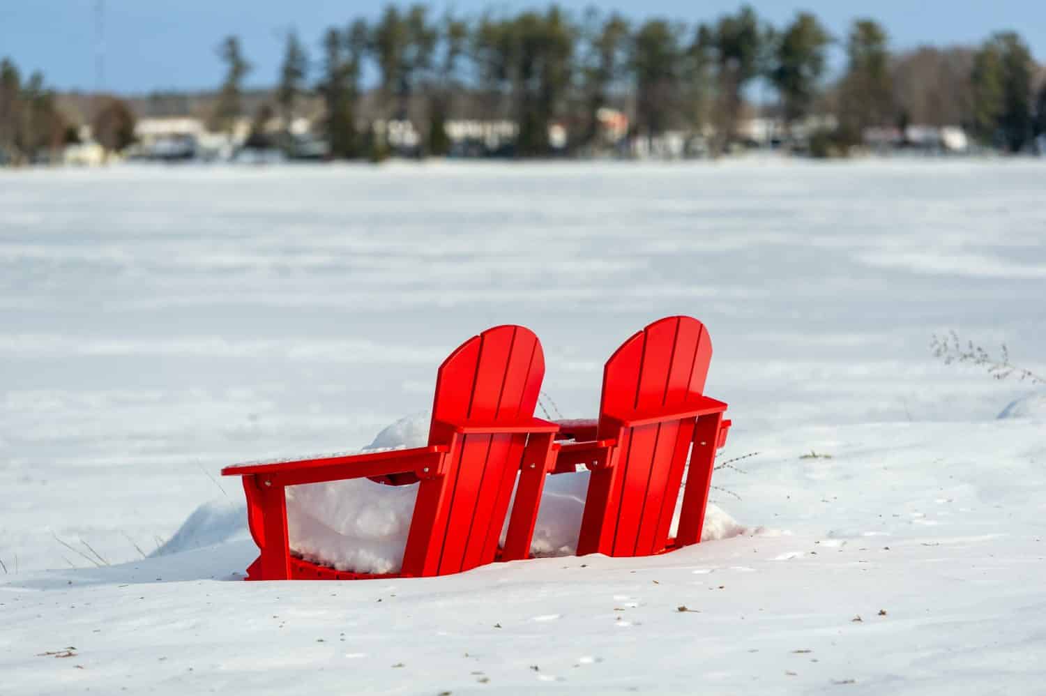 Peaceful winter setting in the Muskoka, Ontario, Canada. Two red Adirondack chairs are covered in a blanket of snow, giving the impression of untouched serenity.