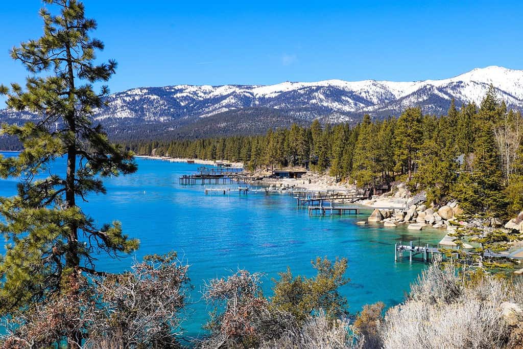 a gorgeous winter landscape with clear blue lake water, snow capped mountain ranges and lush green trees along the banks of the lake and blue sky at Lake Tahoe at Nevada State Park in Incline Village