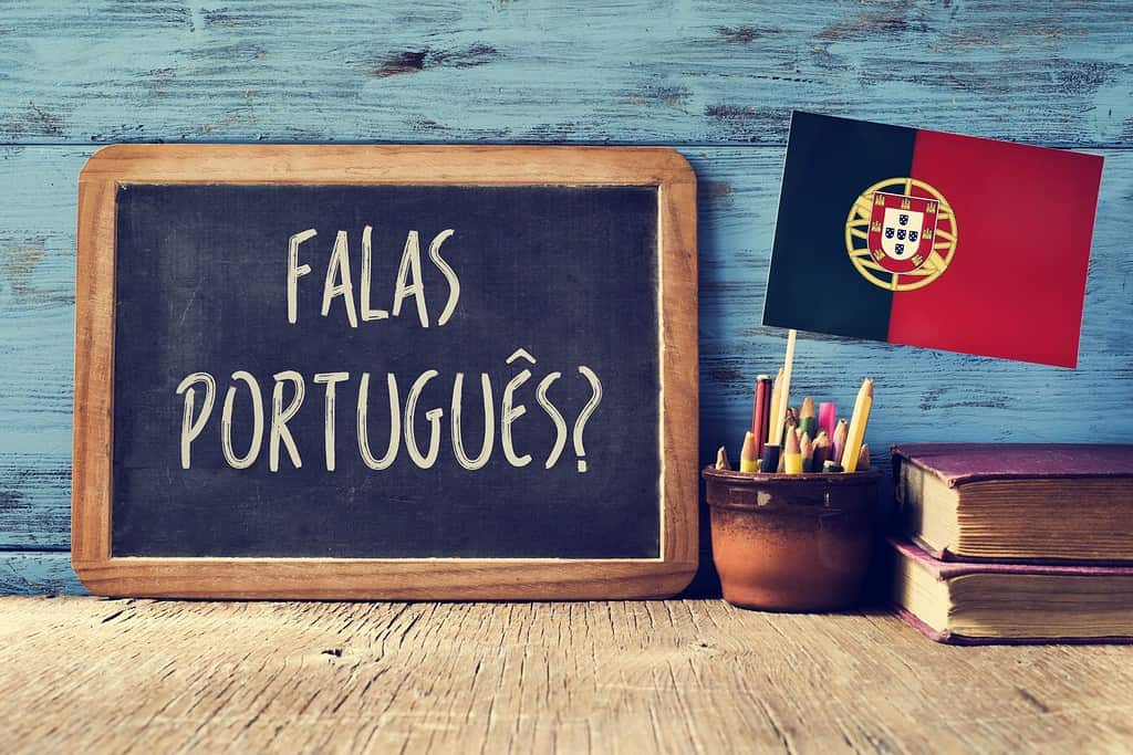 a chalkboard with the question question falas portuges? do you speak Portuguese? written in Portugese, a pot with pencils, some books and the flag of Portugal, on a wooden desk