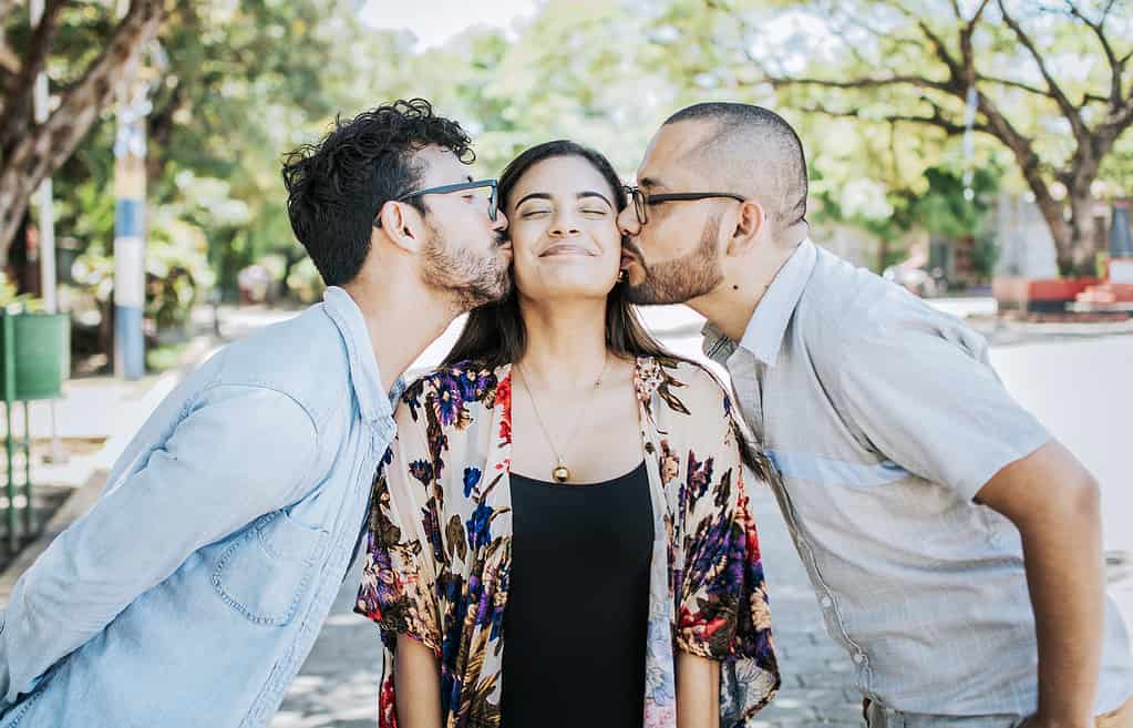 Love triangle concept. Polygamy concept. Two men kissing a girl cheek. Portrait of two guys kissing a girl cheek. Two young men kissing a woman cheek outdoor