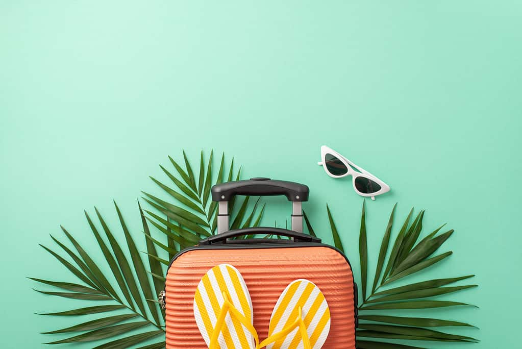 Transport yourself to paradise with top view of a turquoise background showcasing a suitcase, beach essentials, sunglasses, flip-flops and palm leaves. Ideal for travel promotions