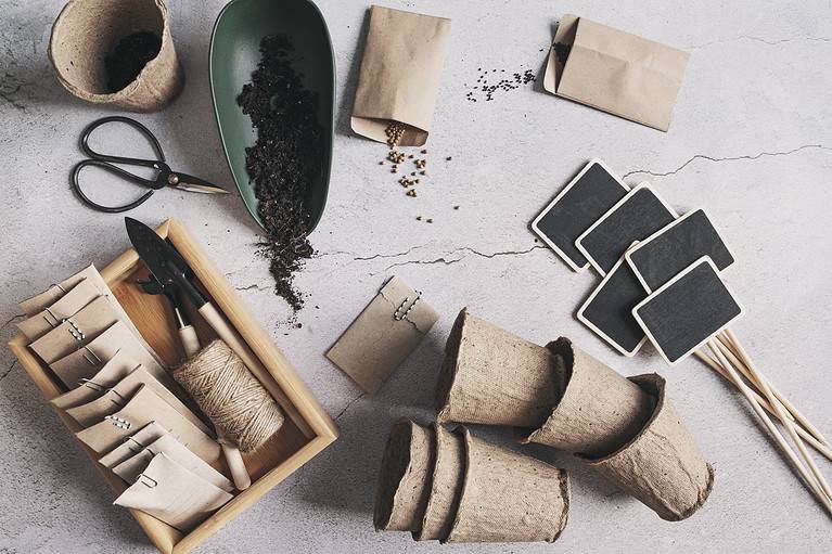 top view of spring seeding process. Tray with soil, paper craft bags with seeds, garden tools and pots on grey background. Seasonal work.
