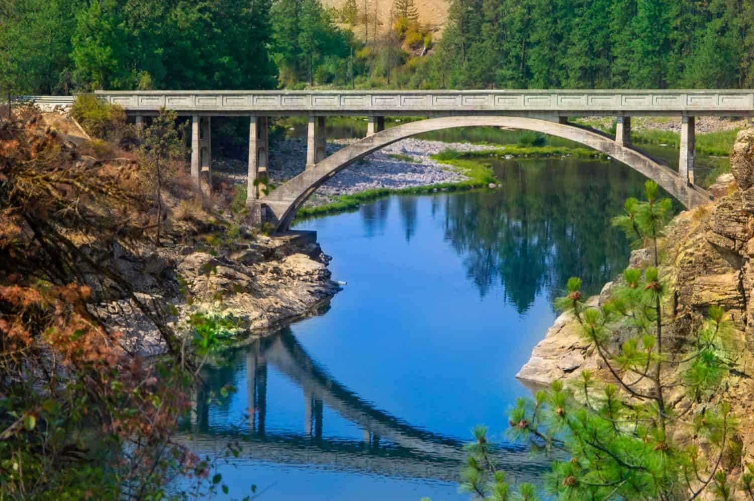 Post Falls, ID—Aug 6, 2018; arched concrete bridge spans Spokane river with blue sky and architecture reflecting off of the water in time exposure with evergreen trees in background.