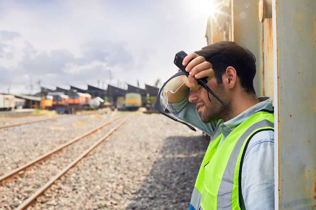 A Railway engineer or Rail transport technician wearing a green safety vest is standing to rest or working outdoors beside a freight train on a hot and sunny day.