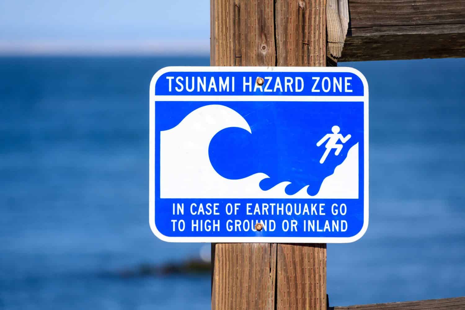 Tsunami Hazard Zone warning sign on ocean coast warns the public about possible danger after an earthquake. Close up. Blurred blue ocean water in background