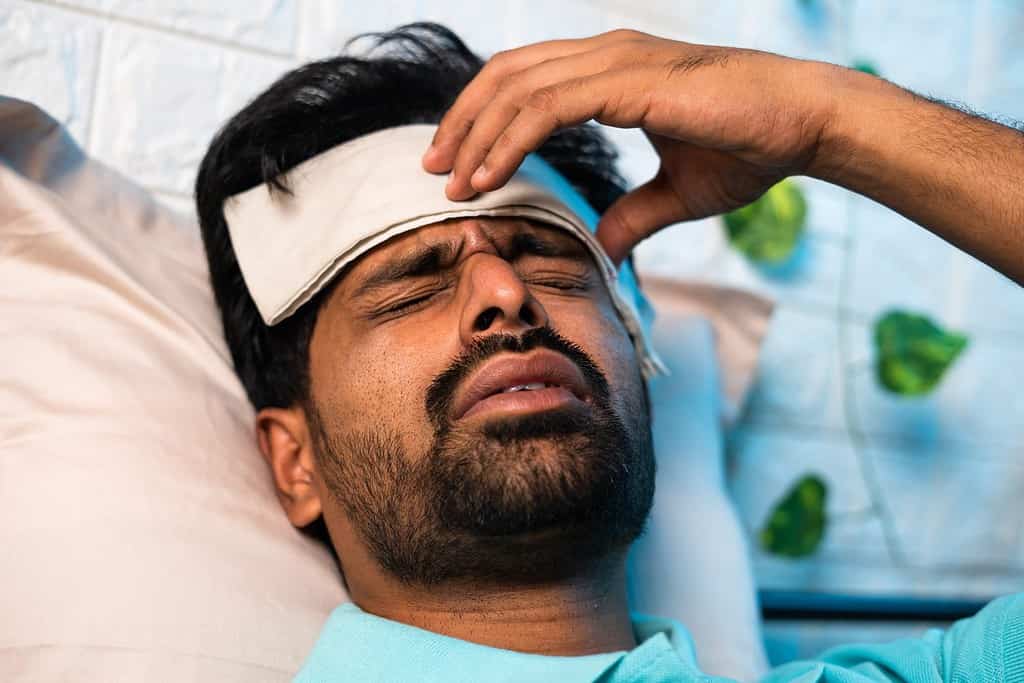 close up head shot of young indian sick man suffering from fever while sleeping at night by placing wet cloth on forehead - concept of healthcare, covid symptoms and illness.