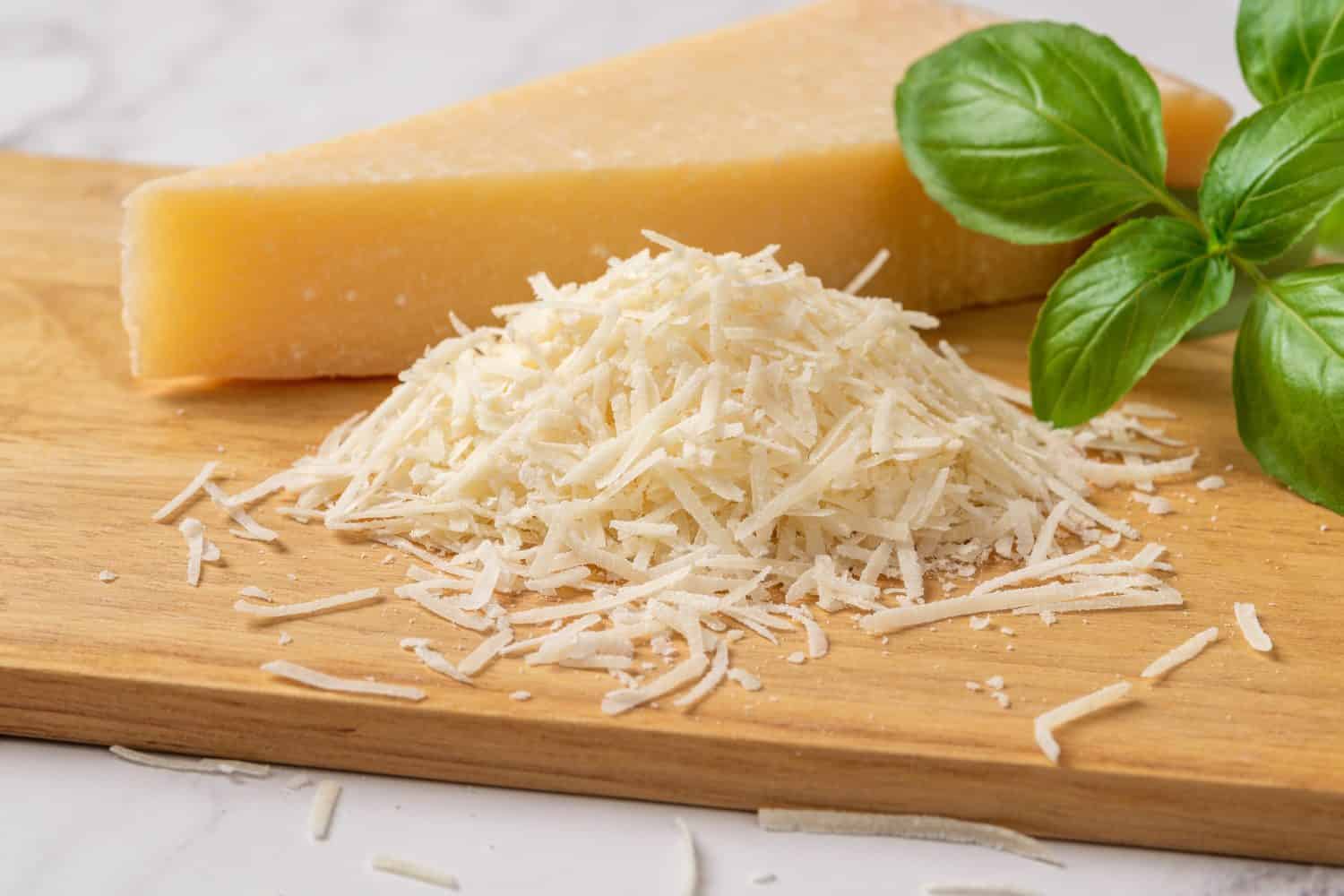 Shredded grana padano cheese on a cutting board. Italian parmesan cheese whole wedge and grated with green basil herb over wooden background. Delicious hard cheese. Dairy product. Front view.