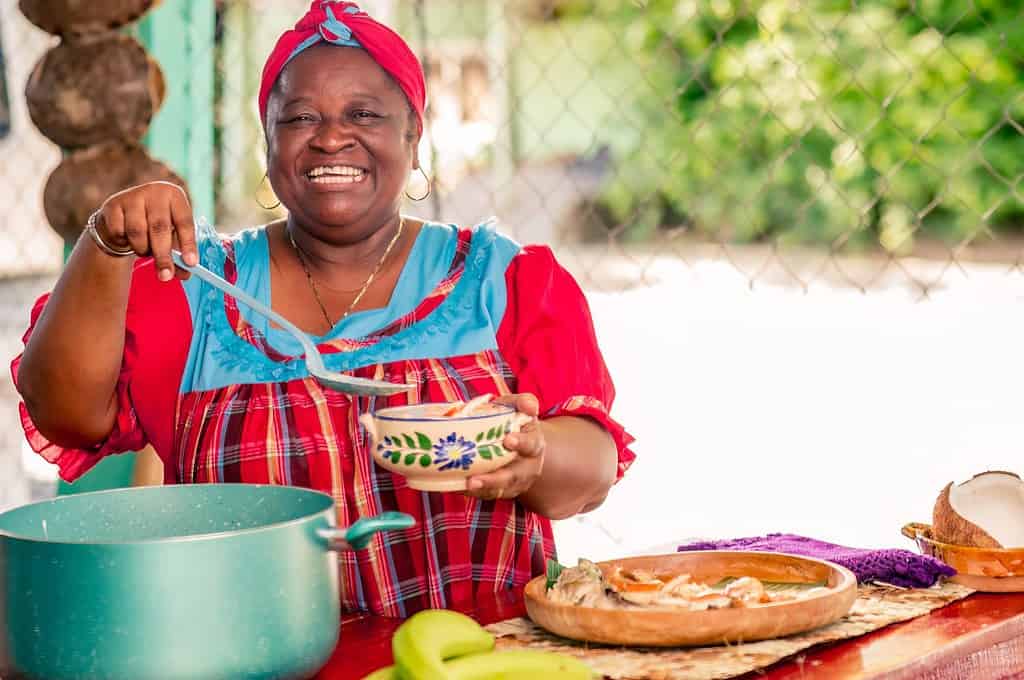 Beautiful black woman of the Garífuna ethnic group smiles at the camera as she puts the hot soup on the plate.