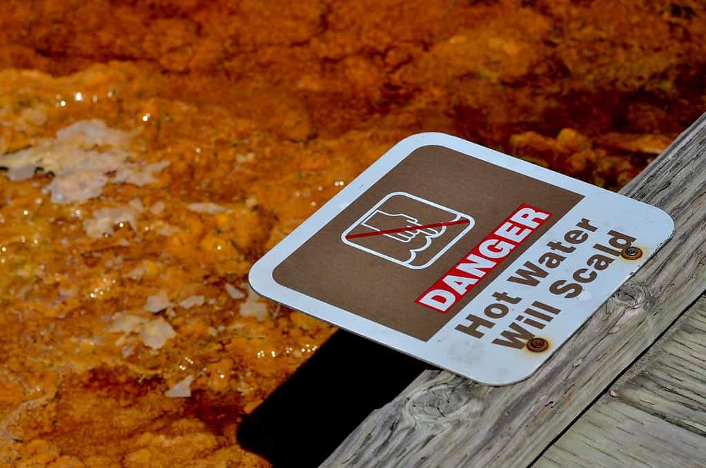 A warning sign reads danger hot water will scald and illustration of a hand dipping into water. The square plate is attached to wooden bridge with orange colored hot spring water in the back.