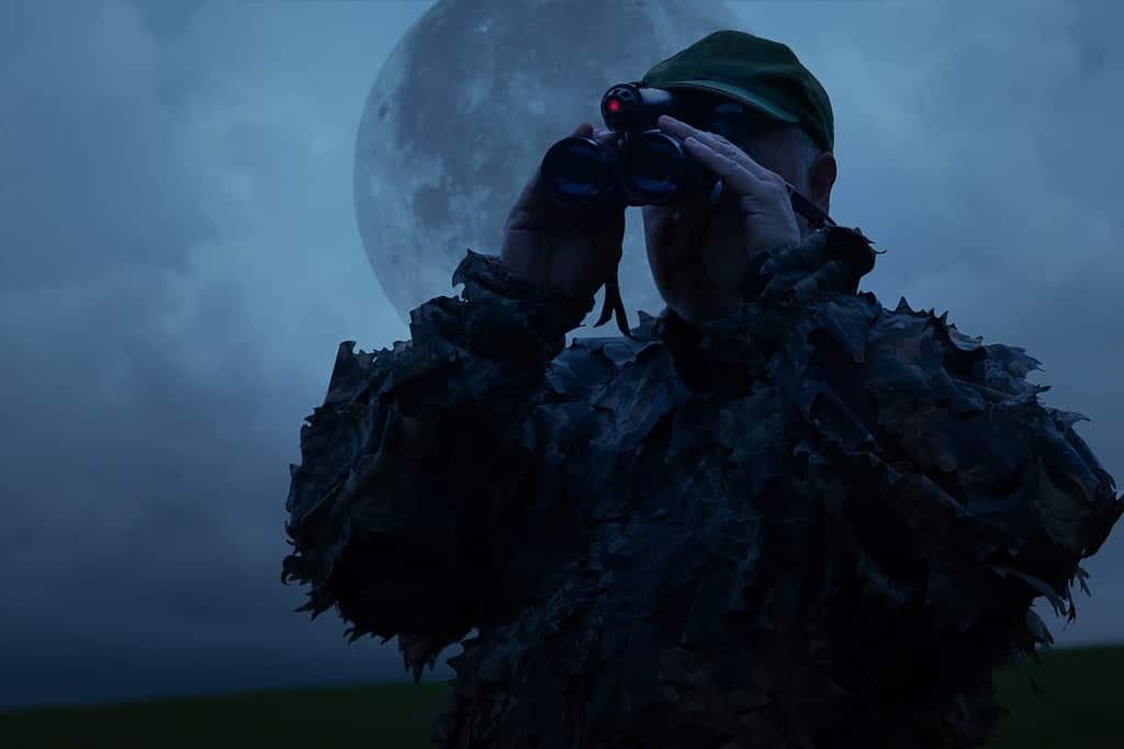 Hunter observes his hunting area with a night vision device under a full moon.
