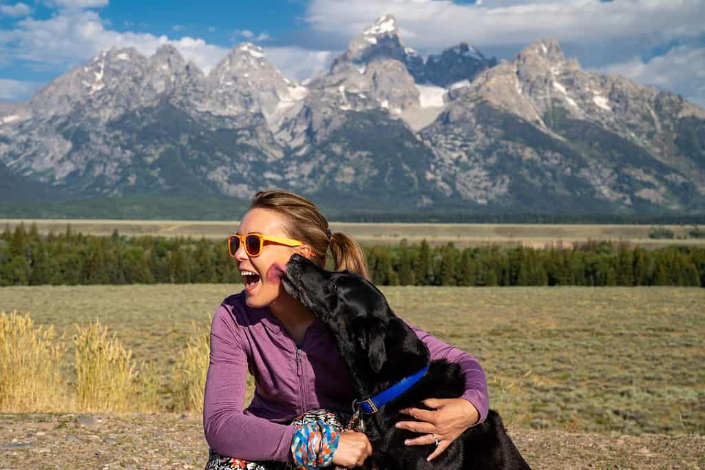 Woman hugs and holds her black labrador retriever dog in front of the Grand Teton National Park mountains in Jackson Wyoming as the dog licks her face
