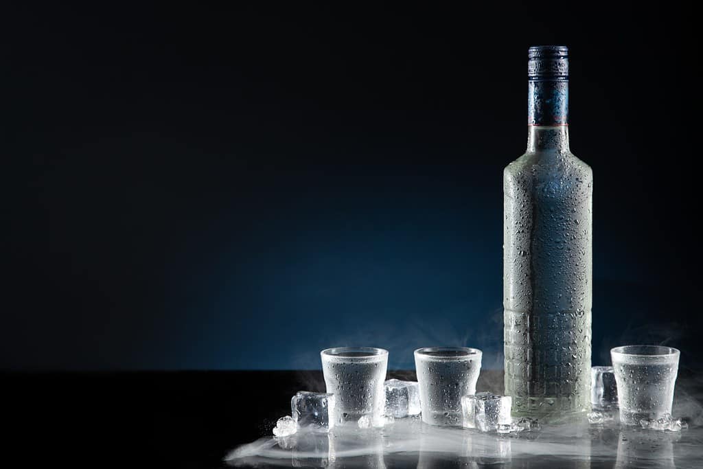 Ice-cold bottle of vodka with shot glasses on dark blue background with copy space.
