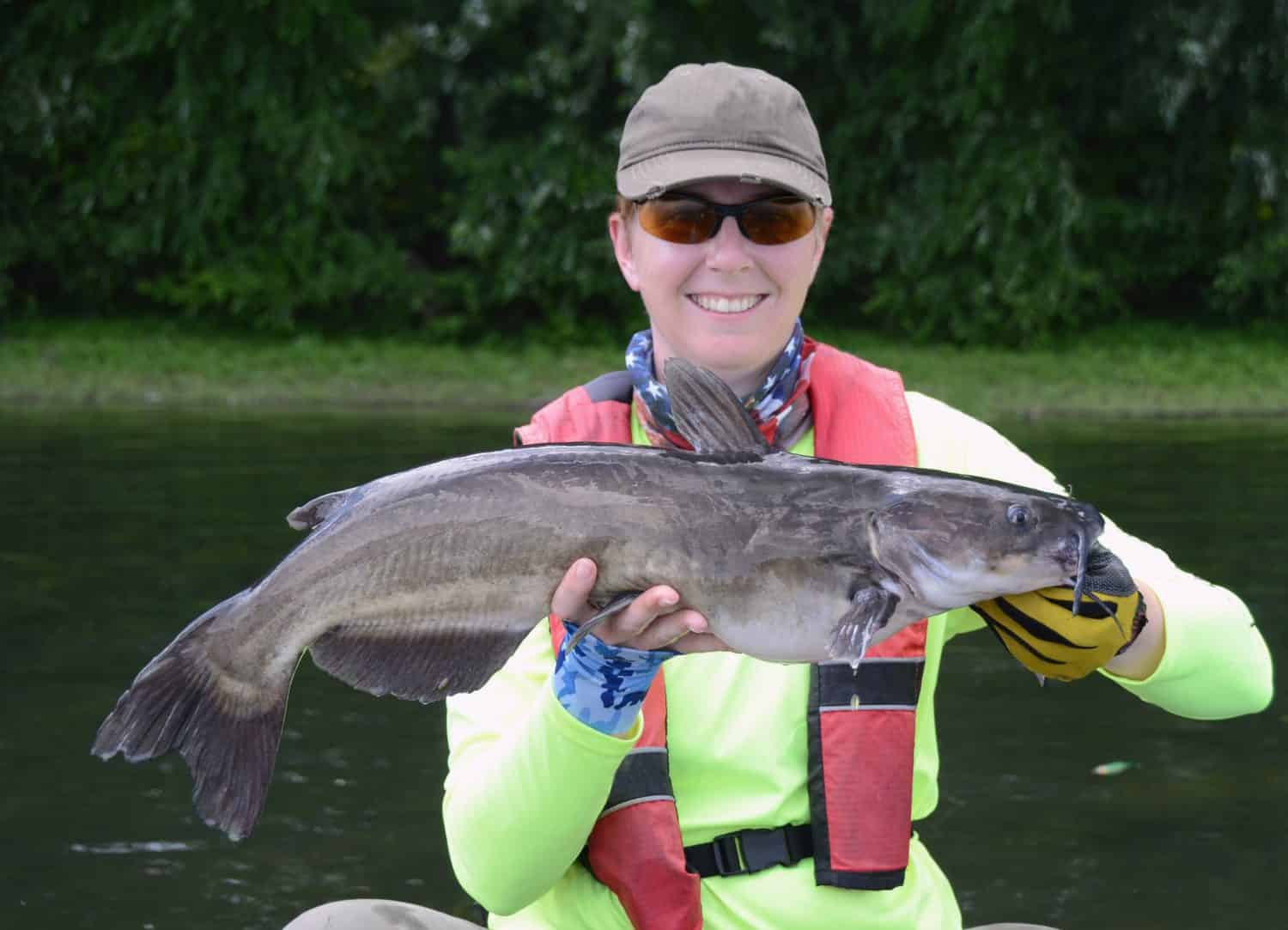 A large dark gray channel catfish fish being held horizontally by smiling short haired woman in a baseball cap and gloves on a bright river on a bright summer day