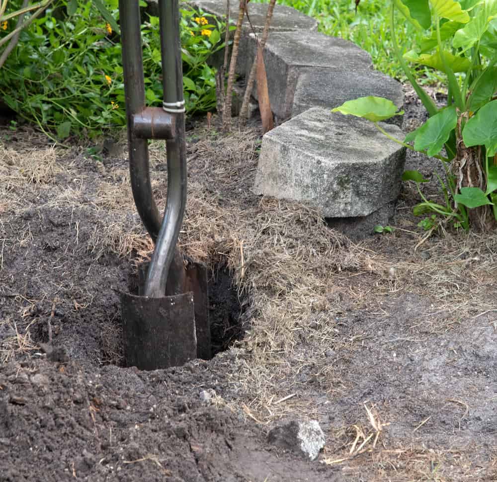 Steel post hole digger with shovel-like blades is being used to create a deep hole for a fence post with concrete bricks and green plants in the background.