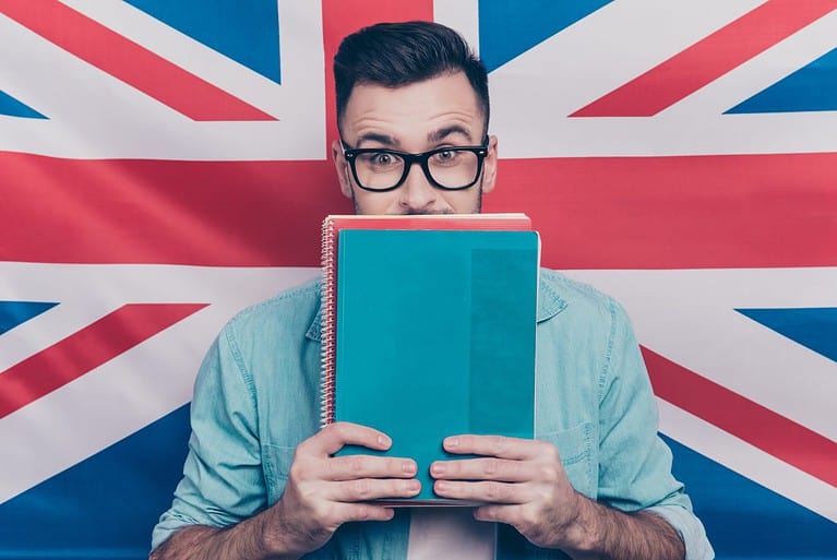 English language learning concept-portrait of excited man holding colorful copy books in hands closing half face with notebooks standing over English flag background