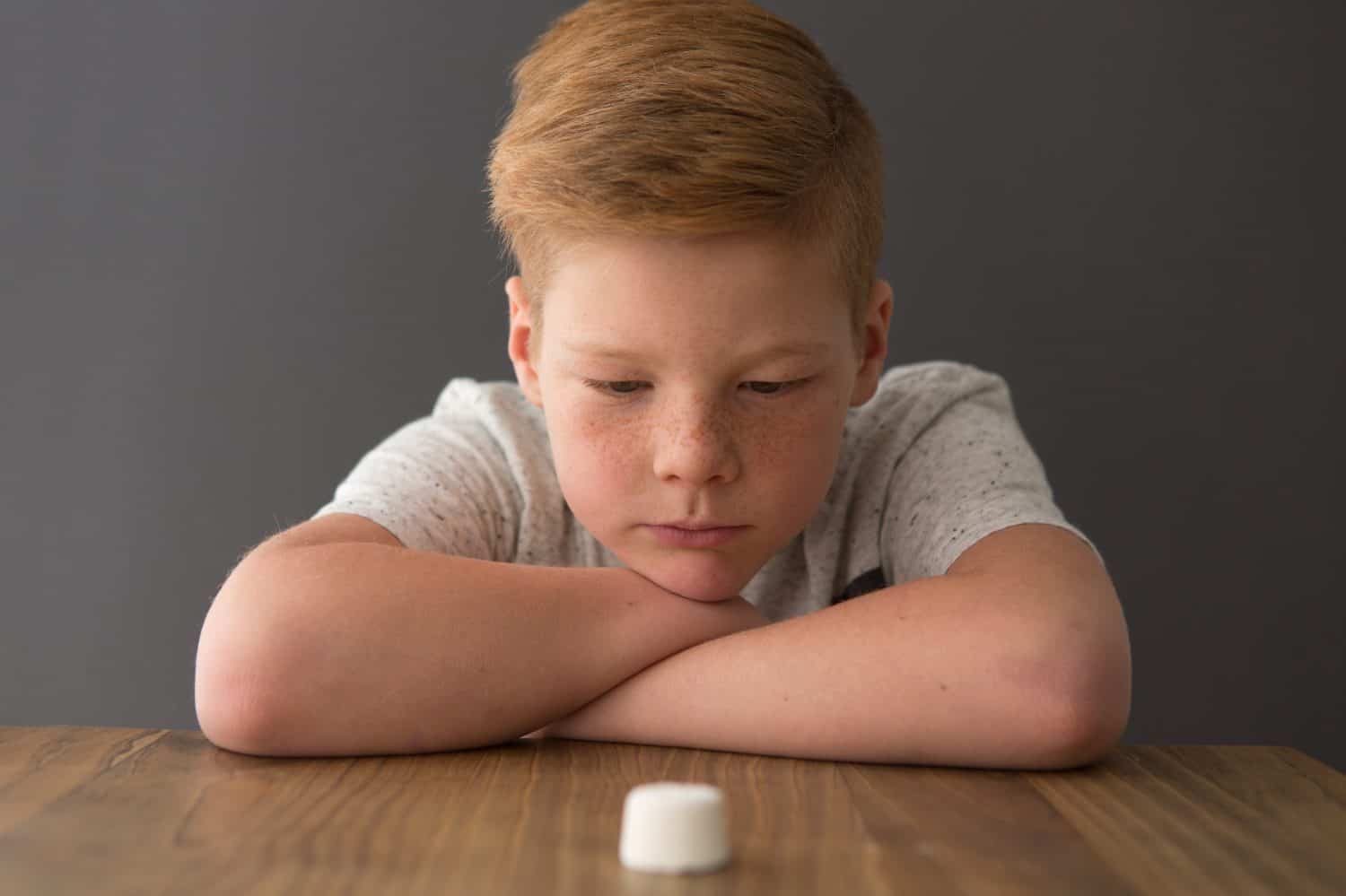 Redhead boy sits at the table opposite a single marshmallow, attempting the marshmallow test