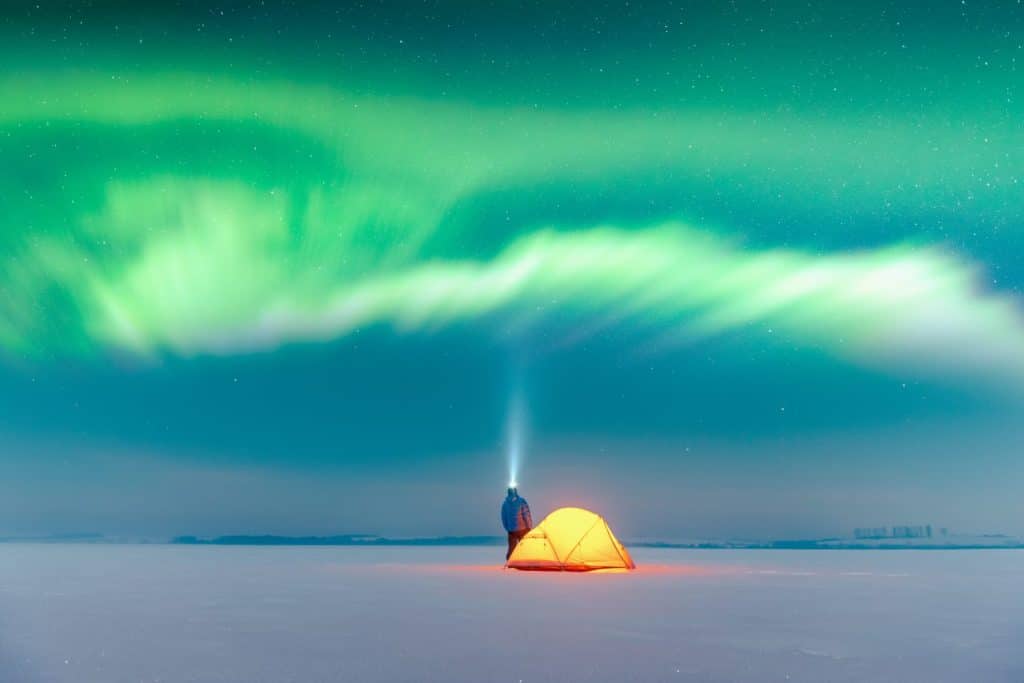 Tourist with flashlight near yellow tent lighted from the inside against the backdrop of incredible starry sky with Aurora borealis. Amazing night landscape. Northern lights in winter field