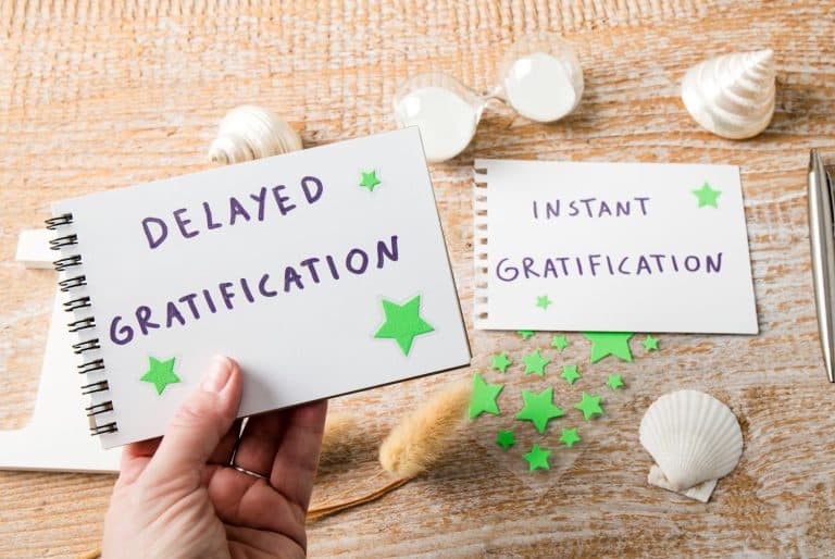 Person hand choosing delayed gratification instead of easy instant gratification. Text on notepad decorated with seashells indoors studio shot.