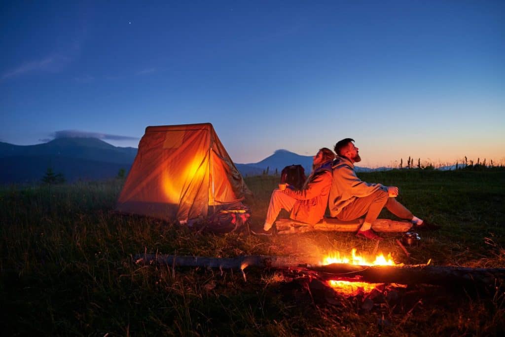 Couple hikers enjoys cozy moment by campfire, sipping hot drinks near glowing tent. Twilight sky and mountain backdrop create serene and romantic atmosphere in picturesque wilderness setting.
