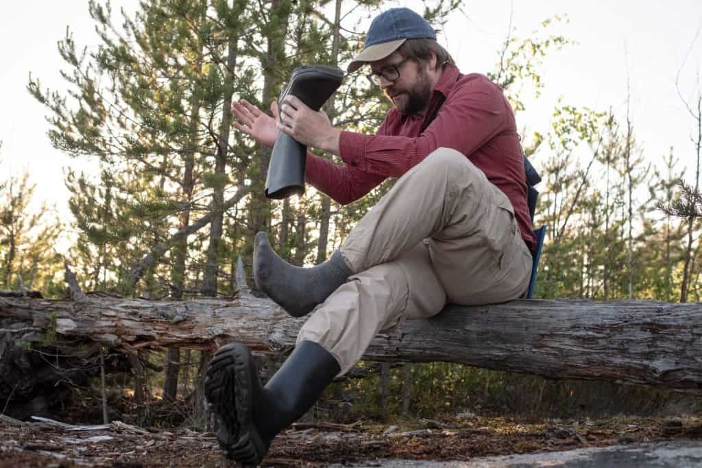 Man in hiking clothes sat on an old log in the woods, took off his rubber boots and shakes out trash. Active lifestyle concept.