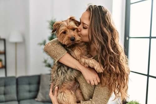 Young beautiful hispanic woman kissing and hugging dog standing at home