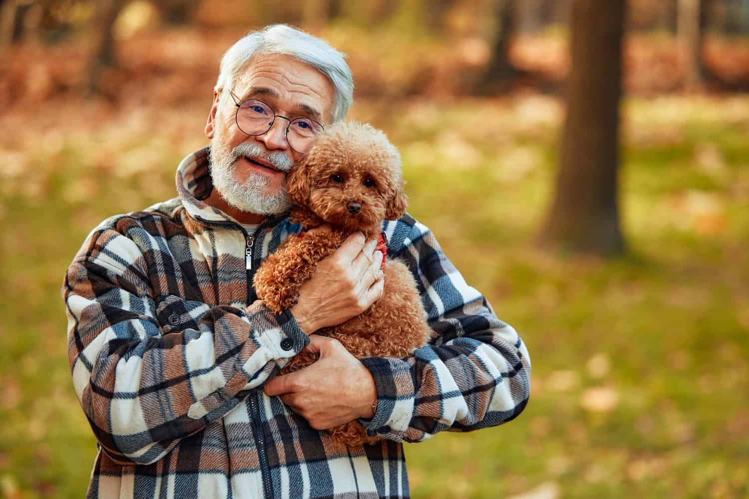 A cheerful elderly man is relishing a delightful moment outdoors while joyfully cradling a fluffy dog, surrounded by a serene and picturesque natural backdrop filled with greenery and tranquility