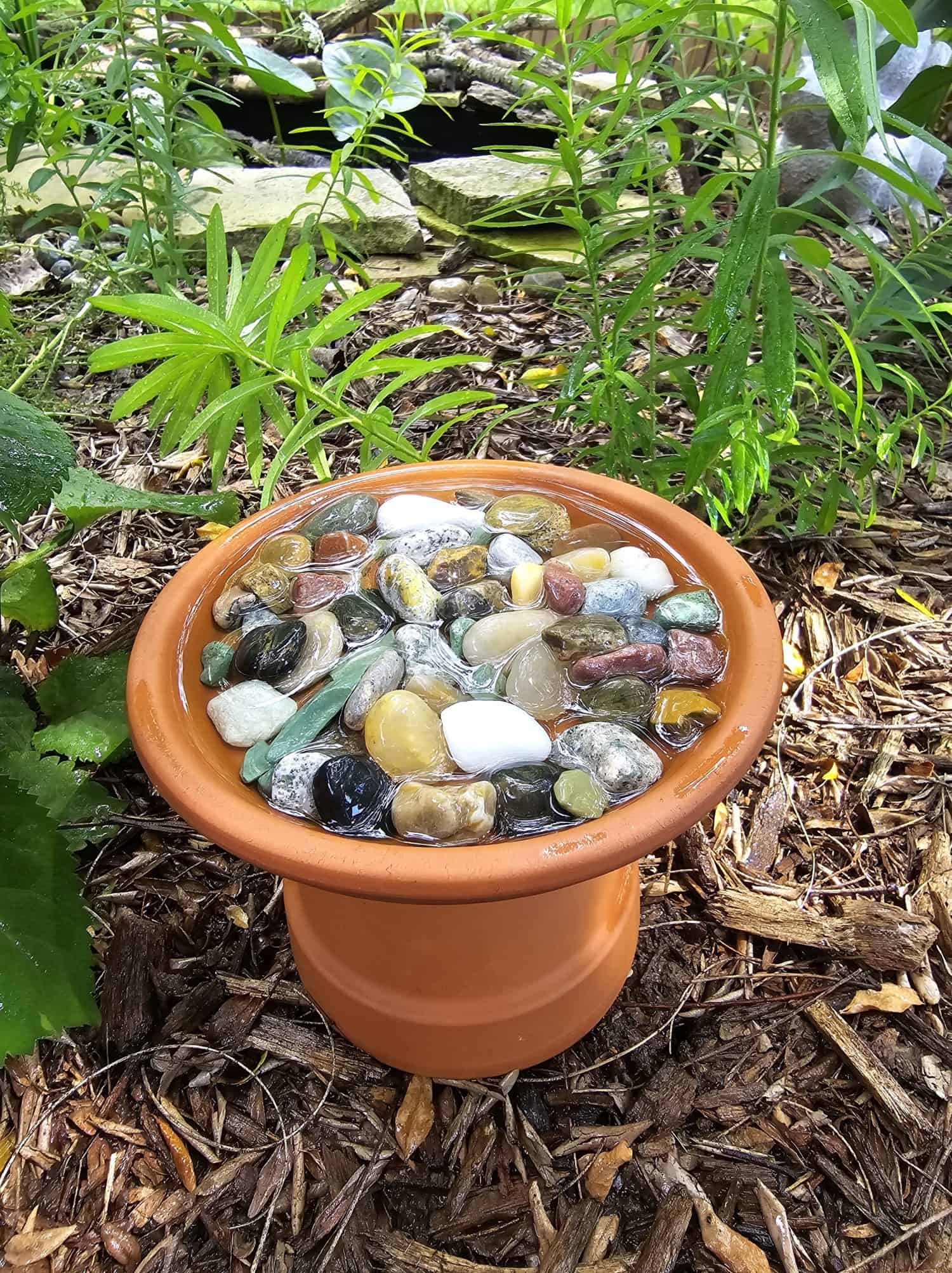 A pollinator water feature made from a terracotta pot with colorful rocks sits on the ground in a garden