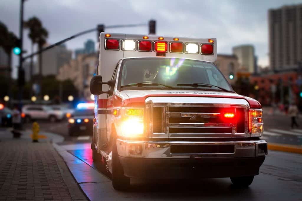 An ambulance with lights activated and a police car behind it in an urban environment