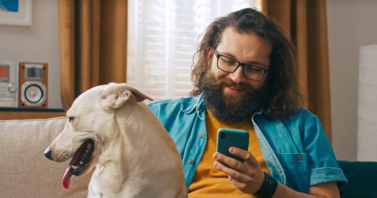 A man sits comfortably on the couch, affectionately petting his dog and reviewing insurance quotes on his smartphone.