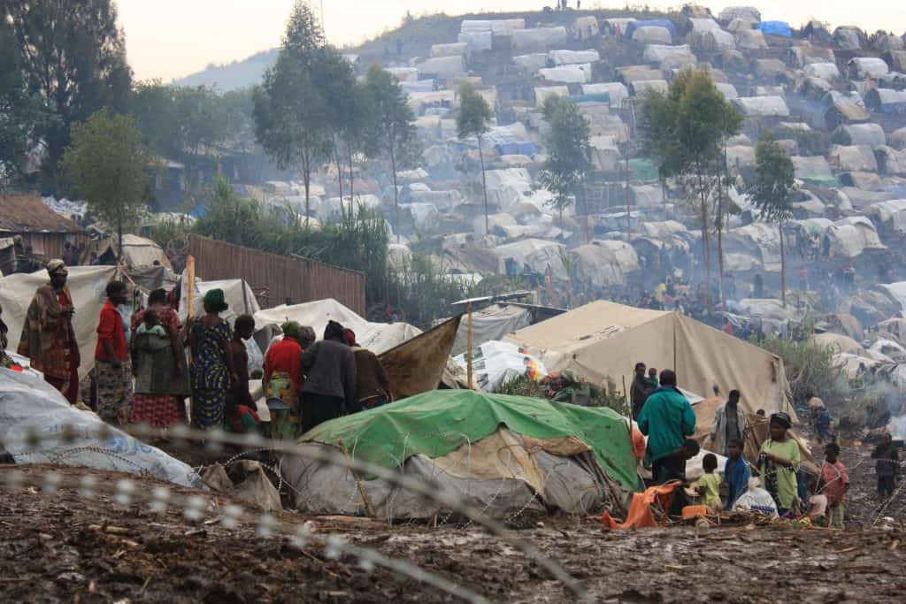 people and tents nestled on a hillside amongst a few trees.