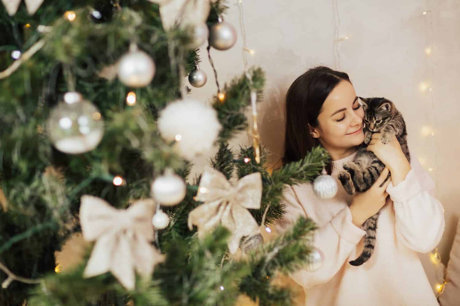 Girl holding in her hands little striped kitten while standing near Christmas tree. She spends the Christmas holidays with her pet. 