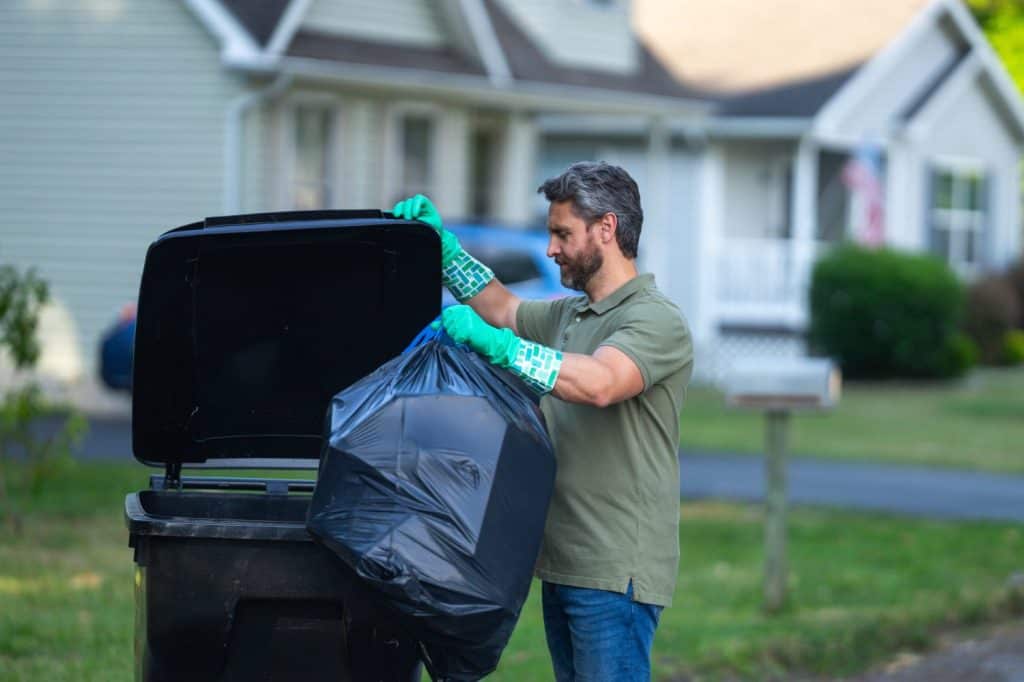 A man pushes a large plastic trash bin for weekly waste disposal schedule. Middle aged man putting out rubbish in garbage bin at the street. Man throwing trash bag into bin outdoors.