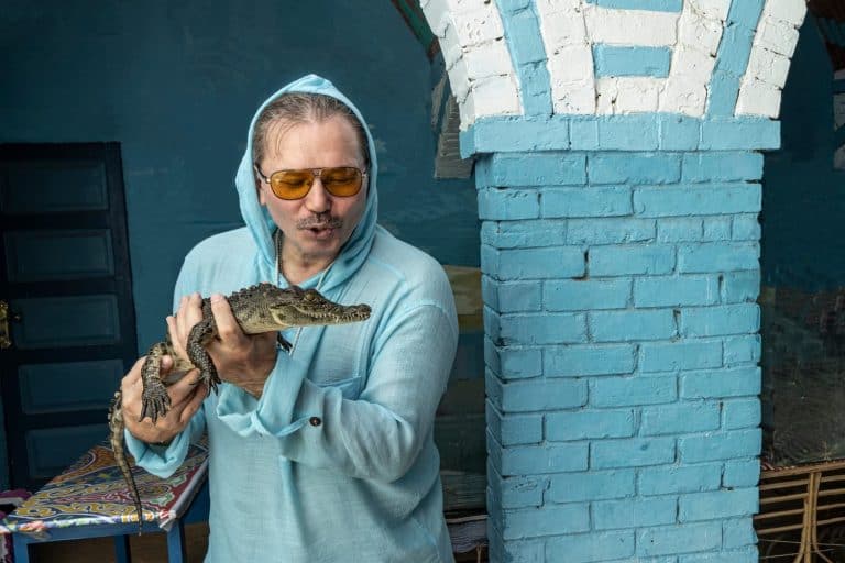 A man holds a small crocodile cub in his hands and smiles. Reptile scales, eyes, teeth, splayed fingers on paws are clearly visible. Next to it is a blue brick column. Egypt