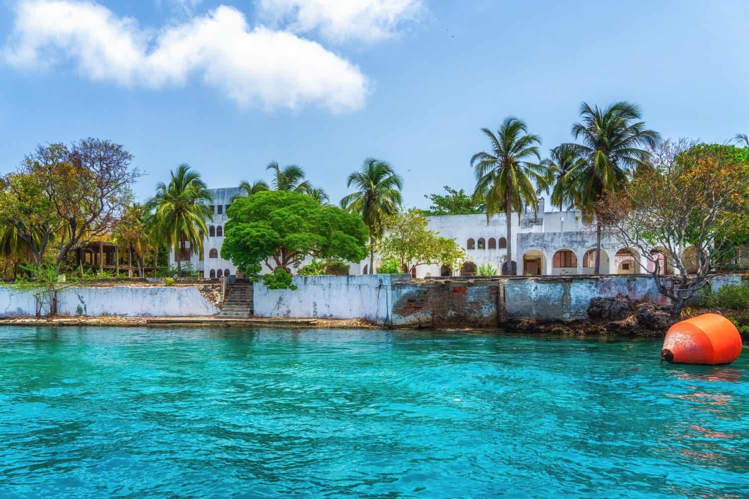 Pablo Escobar abandoned secret island mansion surrounded by tropical trees at Isla Grande. Crumbling white walls visible from the ocean with turquoise water in foreground. Clear blue sky, sunny day.