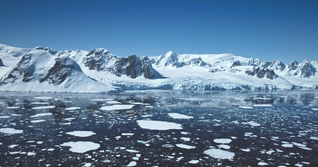 Towering snow-covered mountains in Antarctica. Polar ocean ice floes melting under blue sunny sky. Beautiful South Pole winter horizon . Aerial majestic seascape drone view. Travel exploration