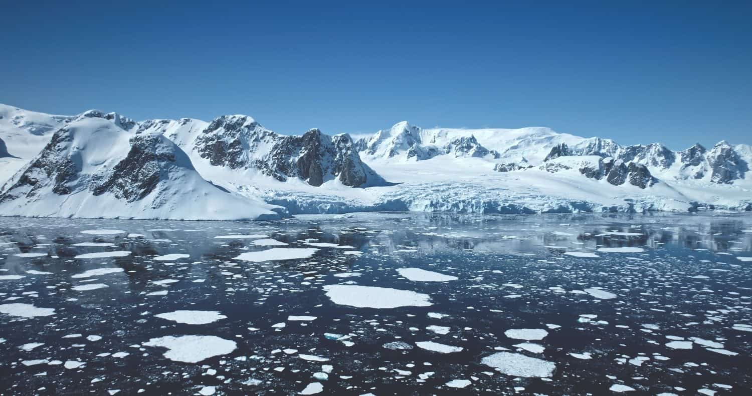 Towering snow-covered mountains in Antarctica. Polar ocean ice floes melting under blue sunny sky. Beautiful South Pole winter horizon . Aerial majestic seascape drone view. Travel exploration