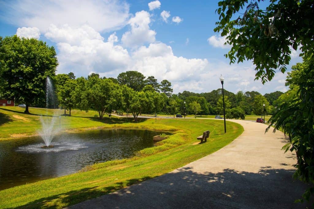 a gorgeous summer landscape in the park with a pond with a water fountain surrounded by lush green trees, grass and plants with blue sky and clouds at Logan Farm Park in Acworth Georgia USA