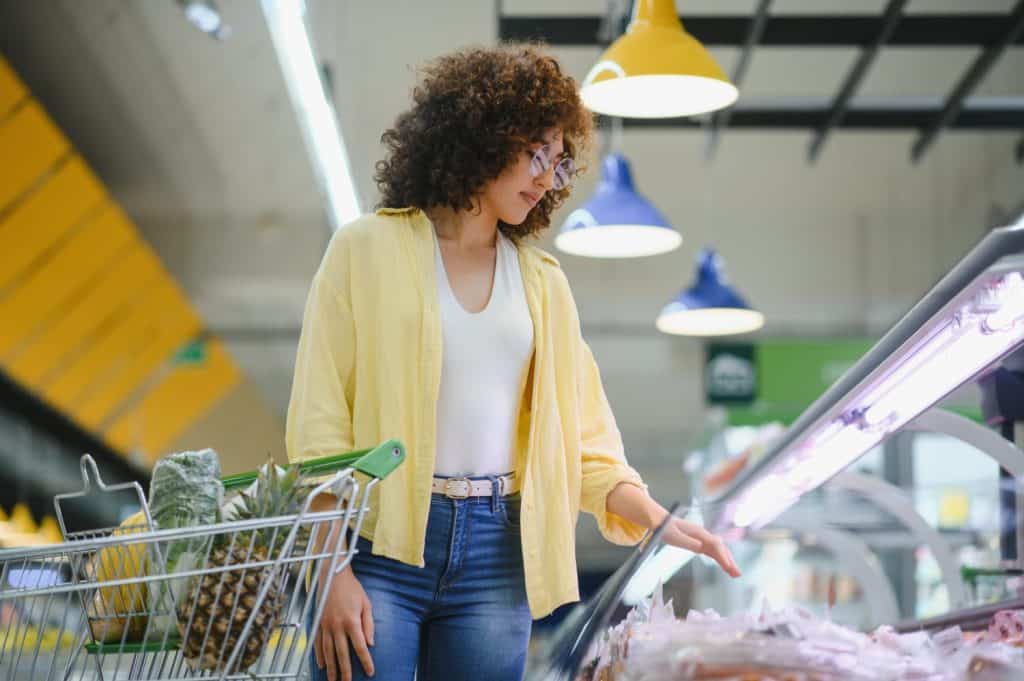 Young woman pushing shopping cart and choosing meat products in refrigerated section of grocery supermarket