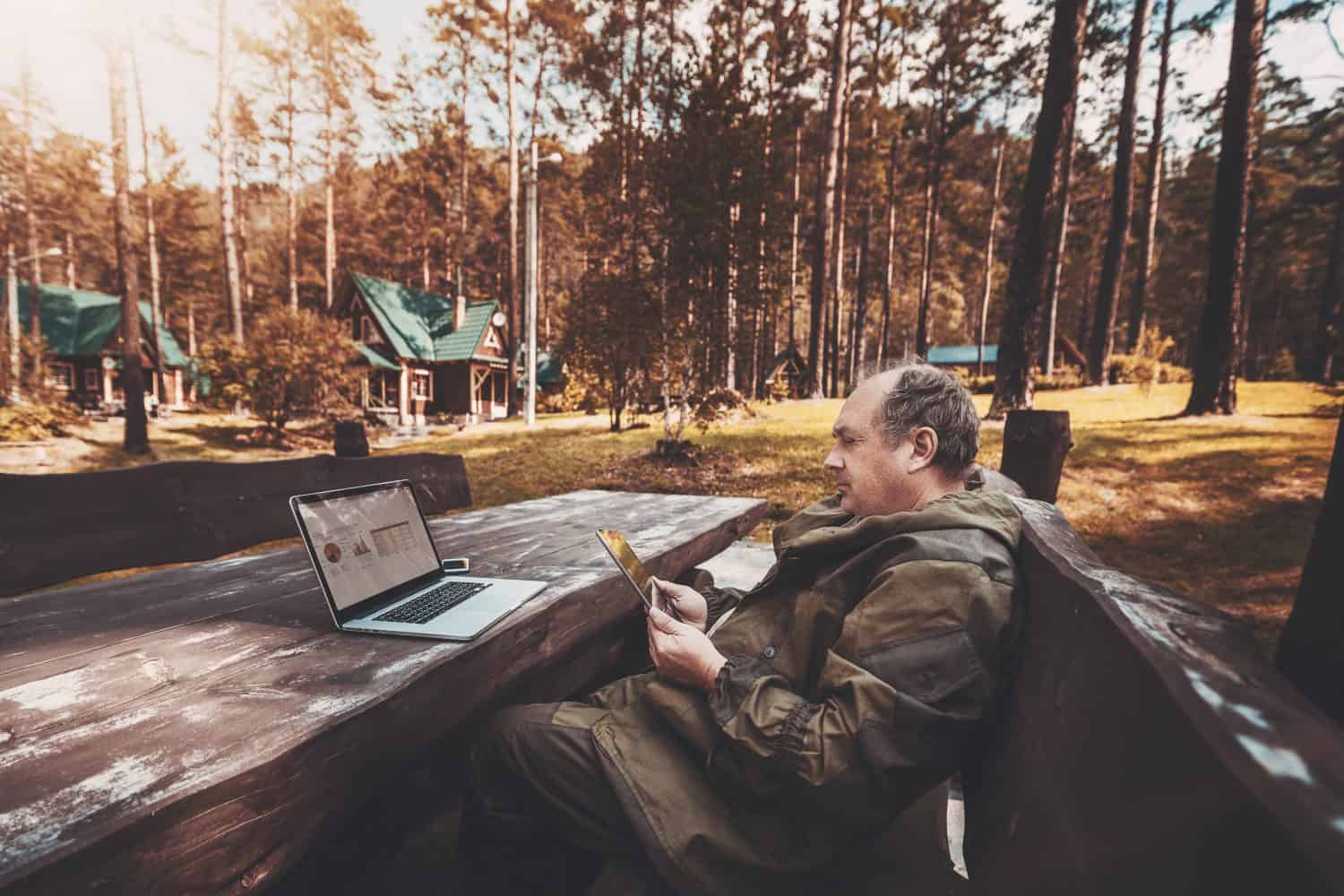 Aged businessman in a dark-green overall is sitting on the wooden bench at the table being on leave and using the digital tablet for video call with his colleagues; laptop with a graph on the table
