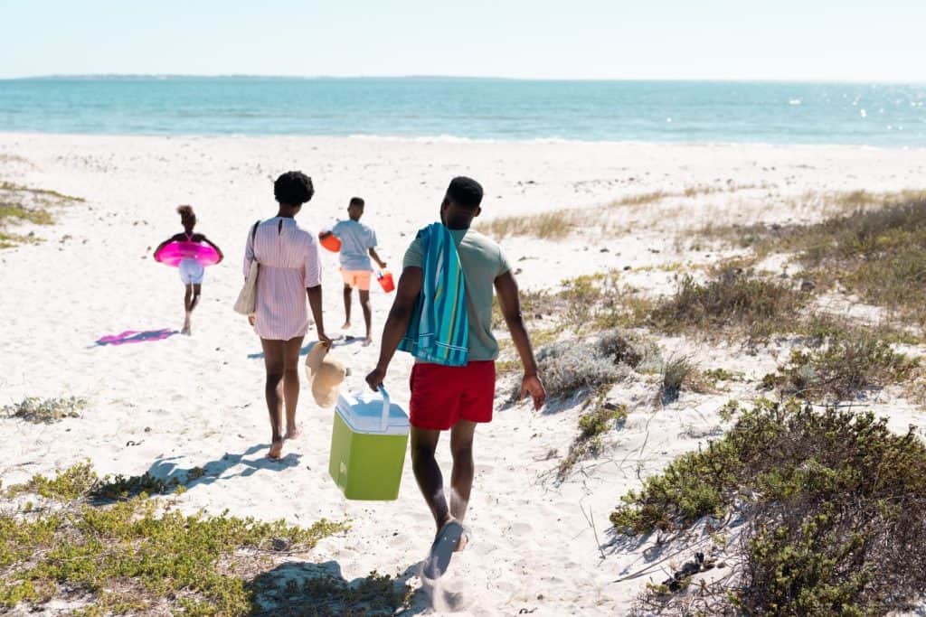 Rear view of african american family with swimming float, cooler walking at beach towards seascape. Copy space, unaltered, parents, together, childhood, picnic, nature, vacation, enjoyment, summer.
